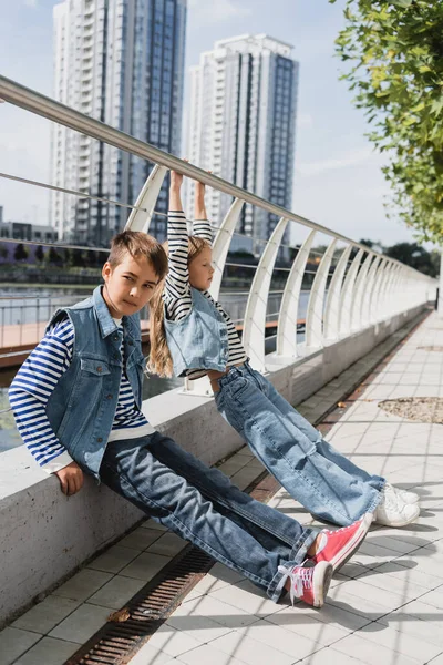Gut gekleidete Kinder in Jeanswesten und Jeans posieren in der Nähe von Metallzaun am Flussufer — Stockfoto