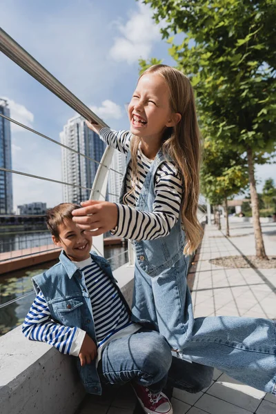 Fröhliches Mädchen in Jeansweste und Jeans neben lachendem Jungen neben Metallzaun am Flussufer — Stockfoto