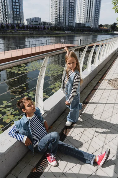 High angle view of well dressed kids in denim vests and jeans posing near metallic fence on embankment of river — Photo de stock