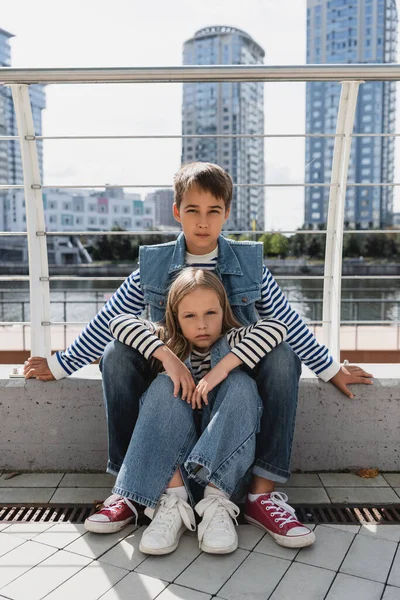 Gut gekleidete Kinder in Jeanswesten und Jeans sitzen am Flussufer neben Metallzaun zusammen — Stockfoto