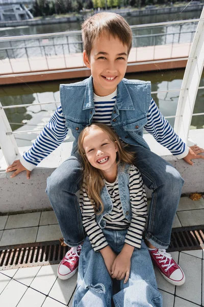 High angle view of well dressed kids in denim vests and jeans smiling next to metallic fence on riverside — Photo de stock