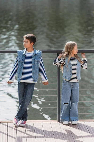 Full length of well dressed kids in denim vests and jeans posing next to metallic fence on river embankment — Stock Photo