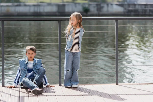 Toute la longueur des enfants bien habillés en gilets en denim et jeans posant à côté de la clôture sur le remblai de la rivière — Photo de stock