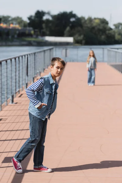 Full length of preteen boy in denim outfit posing near girl standing on river embankment on blurred background - foto de stock
