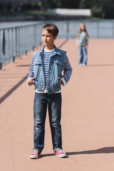 Full length of preteen boy in stylish outfit posing with hands in pockets near blurred girl on river embankment - foto de stock