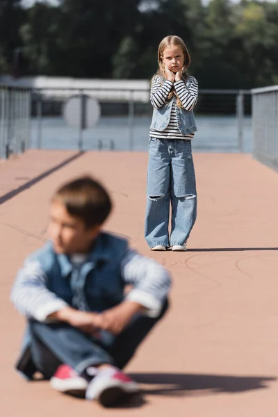 Полная длина девочки-подростка в джинсовой одежде, стоящей на набережной реки рядом с размытым мальчиком на переднем плане — стоковое фото