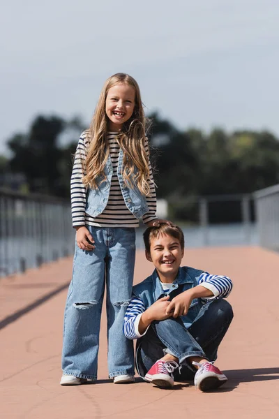 In voller Länge lächelndes Mädchen im Jeans-Outfit, das neben einem glücklichen Jungen am Ufer des Flusses steht — Stockfoto