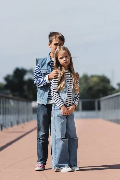 Full length of preteen boy and girl in denim clothes standing on riverside embankment — стоковое фото