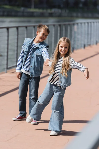 In voller Länge glückliche Kinder in Jeanskleidung, die sich Händchen haltend am Ufer des Flusses amüsieren — Stockfoto