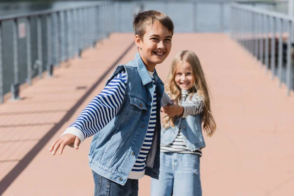 Happy kids in denim clothes holding hands while having fun on riverside embankment — Fotografia de Stock