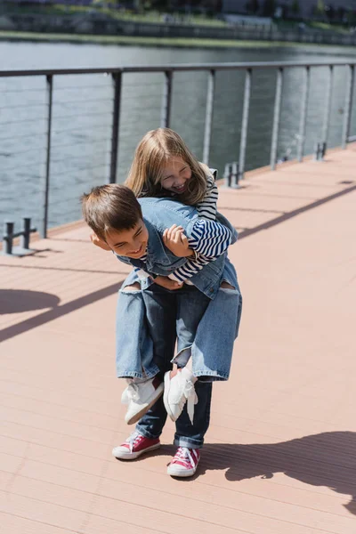 Happy preteen boy in denim outfit piggybacking smiling girl on river embankment — Stock Photo