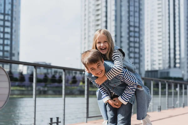 Glücklicher preteen Junge im Jeans-Outfit huckepack Mädchen auf Flussufer — Stockfoto