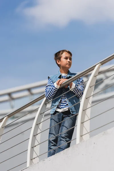 Low angle view of preteen boy in trendy denim outfit posing near metallic fence on embankment — Foto stock