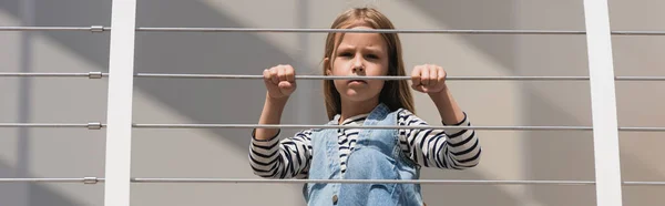 Low angle view of stylish kid in denim outfit looking at camera near metallic fence, banner — Fotografia de Stock