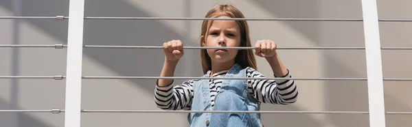 Low angle view of stylish kid in denim outfit looking at camera near metallic fence, banner — Stock Photo