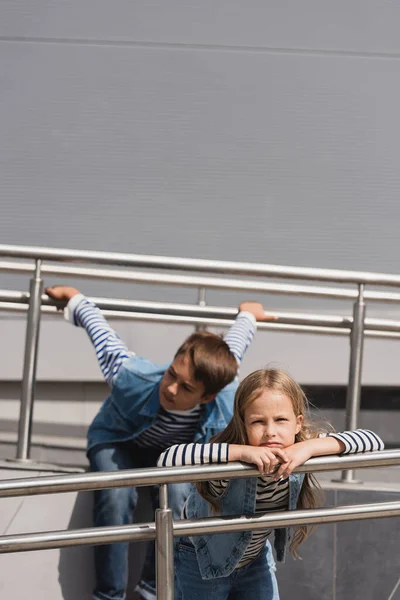 Well dressed girl in casual denim attire posing near metallic handrails next to boy on blurred background — стоковое фото