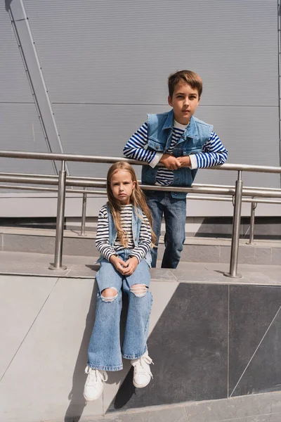 Stylish kids in casual denim attire posing near metallic handrails next to building — Photo de stock