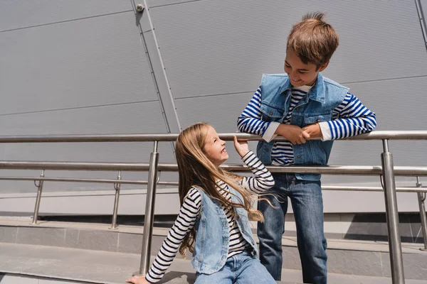 Happy and well dressed kids in casual denim attire posing near metallic handrails next to building — Stockfoto
