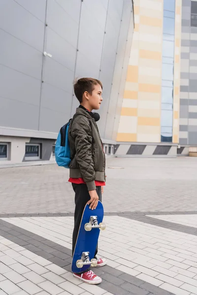 Side view of preteen boy in bomber jacket holding penny board while standing near mall — Photo de stock