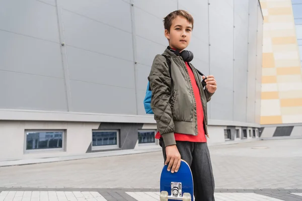 Preteen boy in bomber jacket holding penny board while standing near mall — Photo de stock