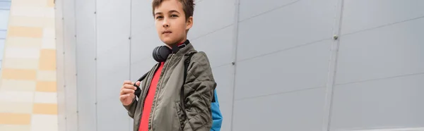 Preteen boy in wireless headphones standing with backpack near mall, banner — Stock Photo