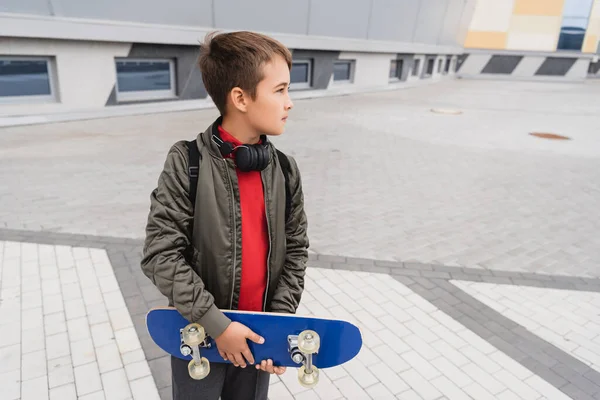 Niño preadolescente en chaqueta de bombardero y auriculares inalámbricos que sostienen penny board mientras está de pie cerca del edificio del centro comercial - foto de stock