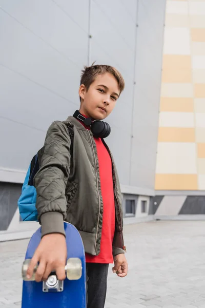 Stylish boy in bomber jacket and wireless headphones holding penny board while standing near mall — Stock Photo