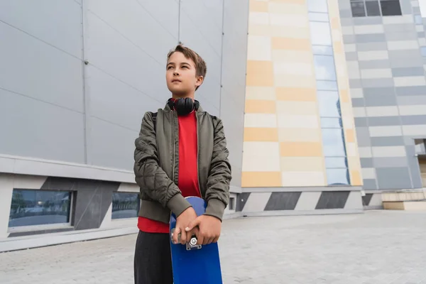 Preteen boy in bomber jacket and wireless headphones holding penny board while looking away near mall — Stock Photo