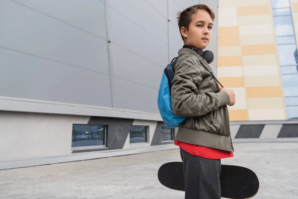Хлопчик у бездротових навушниках стоїть з рюкзаком і тримає дошку копійки біля будівлі торгового центру — стокове фото