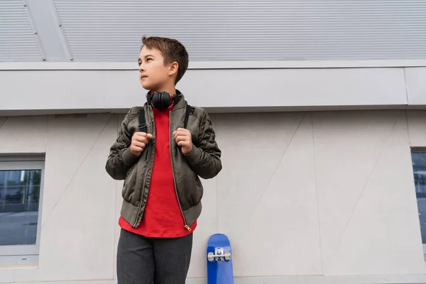 Preteen boy in bomber jacket and wireless headphones standing with backpack near penny board - foto de stock