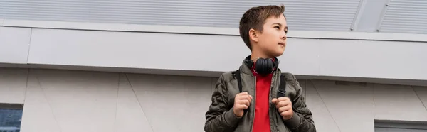 Preteen boy in bomber jacket and wireless headphones holding backpack while standing near mall, banner — Foto stock