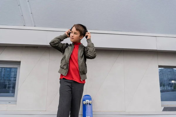 Preteen boy in bomber jacket wearing wireless headphones near penny board while standing near mall — Stock Photo