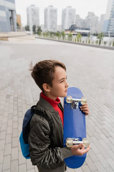 Vista lateral del niño preadolescente en la chaqueta de bomber de moda de pie con la mochila mientras sostiene penny board - foto de stock