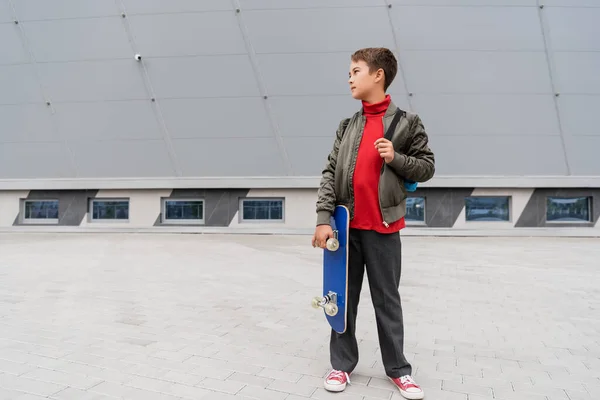 Comprimento total do menino pré-adolescente na jaqueta de bombardeiro na moda segurando penny board perto do prédio do shopping — Fotografia de Stock