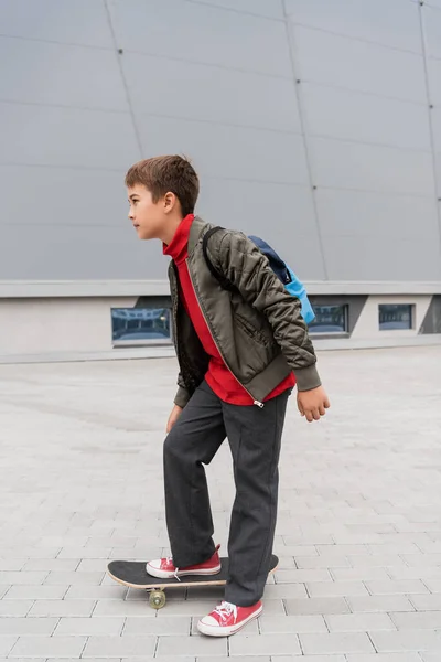 Full length of preteen boy in trendy bomber jacket riding penny board near mall - foto de stock