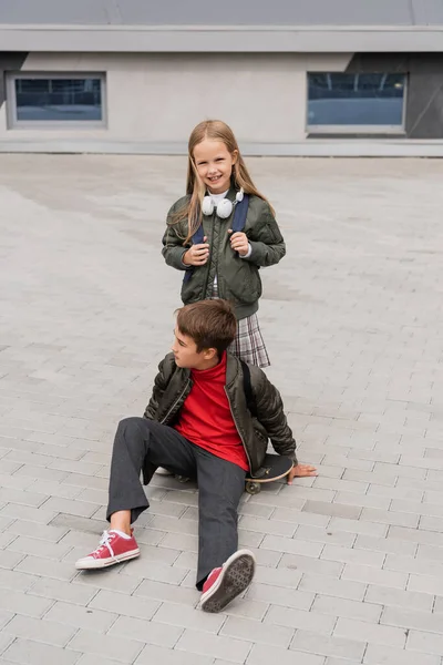 Longitud completa de niño preadolescente en chaqueta de bomber de moda sentado en el tablero de penique cerca de chica alegre - foto de stock