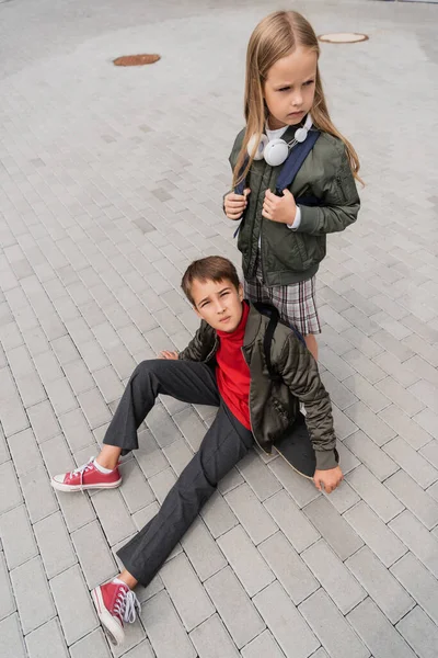 Vista aérea de menino pré-adolescente na jaqueta de bombardeiro na moda sentado na placa penny perto da menina em fones de ouvido sem fio — Fotografia de Stock
