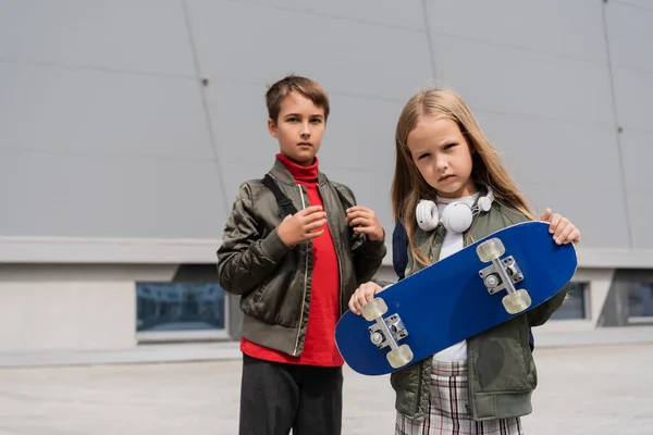 Preteen girl in wireless headphones holding penny board while standing with boy near building of mall — Stock Photo