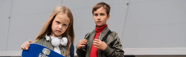 Дівчина в бездротових навушниках тримає дошку пенні, стоячи з хлопчиком біля торгового центру, банер — стокове фото