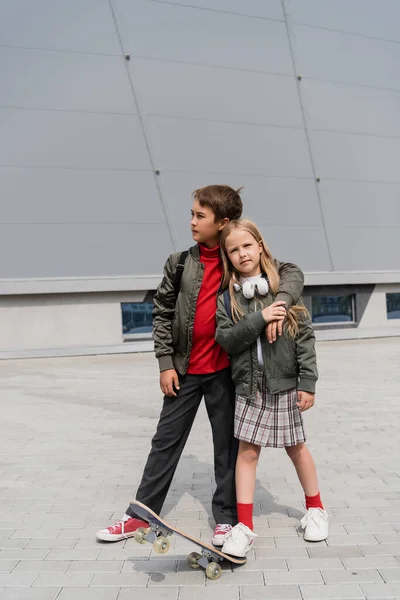 Full length of preteen boy in trendy bomber jacket hugging girl in skirt while standing near penny board — Stockfoto
