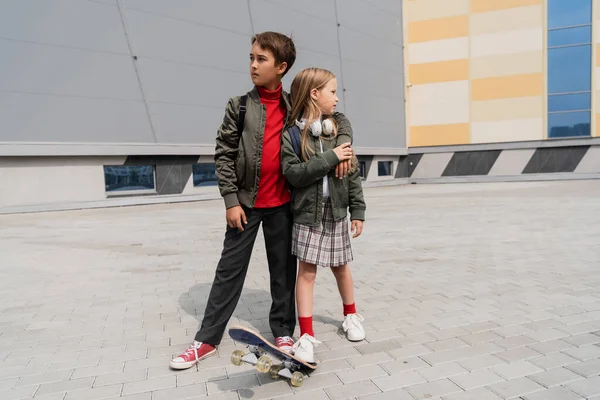 Full length of preteen boy in stylish bomber jacket hugging girl in skirt while standing near penny board — Photo de stock