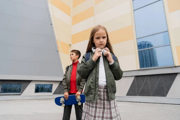 Preteen girl in wireless headphones standing with stylish boy holding penny board on blurred background — Fotografia de Stock