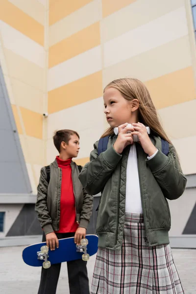 Niña preadolescente en auriculares inalámbricos de pie cerca de un niño con estilo que sostiene el tablero de penique sobre fondo borroso - foto de stock