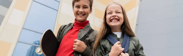 Niño preadolescente feliz sosteniendo penny board mientras está de pie con chica cerca del centro comercial, pancarta - foto de stock