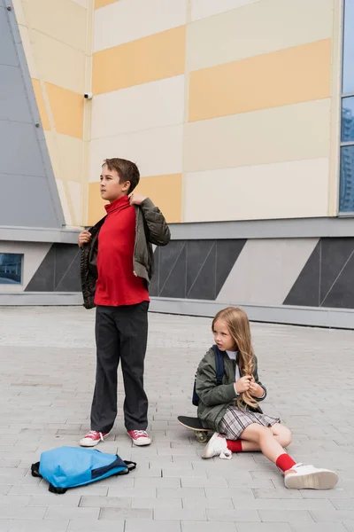 Preteen girl in skirt sitting on penny board next to stylish boy wearing bomber jacket near mall - foto de stock