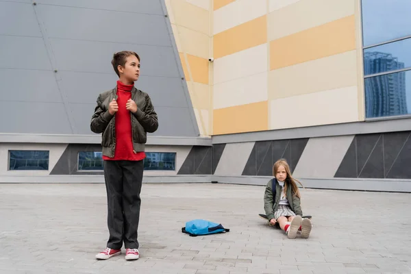 Preteen girl in skirt sitting on penny board near stylish boy in trendy bomber jacket next to mall — стоковое фото
