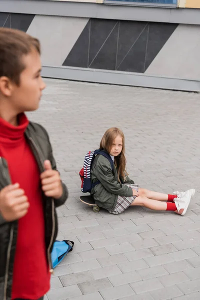 Preteen girl in skirt sitting on penny board near stylish boy on blurred foreground - foto de stock