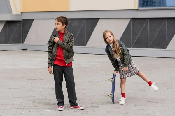 Longitud completa de la niña preadolescente en falda sosteniendo penny board cerca de niño en chaqueta de bombardero, mientras que de pie junto al centro comercial - foto de stock