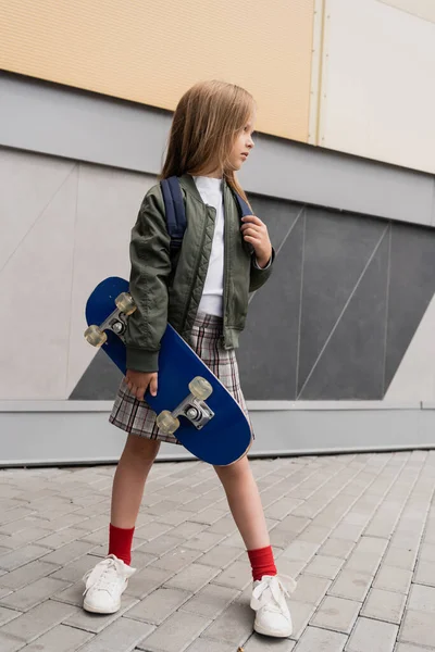 Full length of stylish preteen girl in bomber jacket holding penny board standing near mall - foto de stock