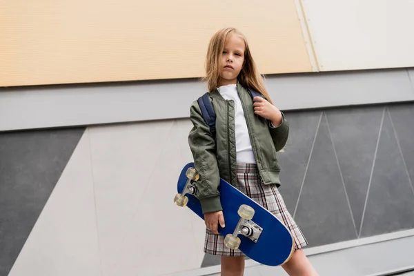 Vista de ángulo bajo de la niña preadolescente en la chaqueta de bombardero de moda que sostiene penny board cerca del centro comercial - foto de stock